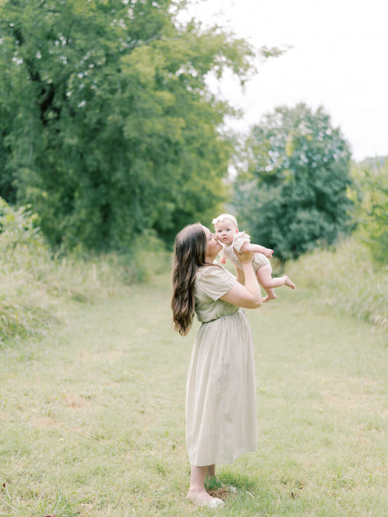 Mother standing in a grassy field with trees, holding up her 6 month old daughter to nuzzle her.