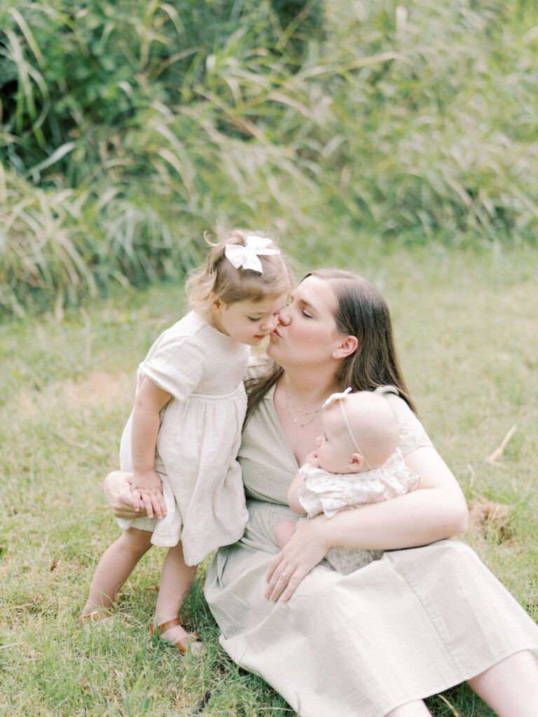 Mother in a green dress sitting in the grass holding her 6 month old daughter while pulling her 2 year old daughter in for a kiss.