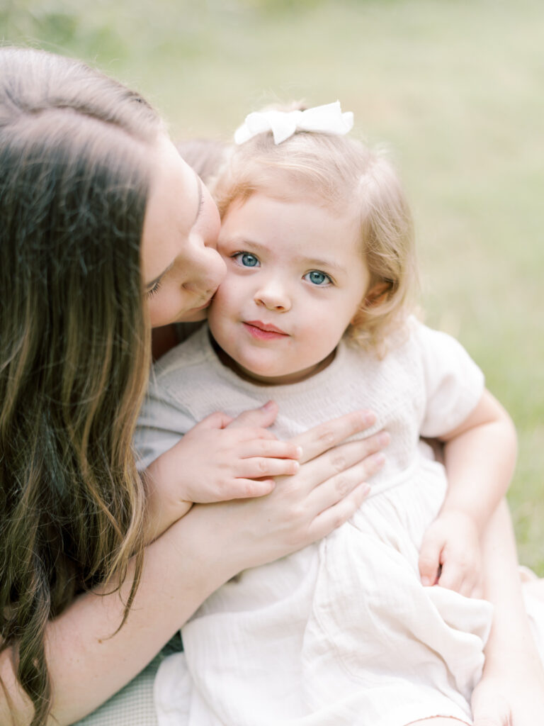 2 year old brunette daughter sitting in her mother's lap while being kissed by her mother.