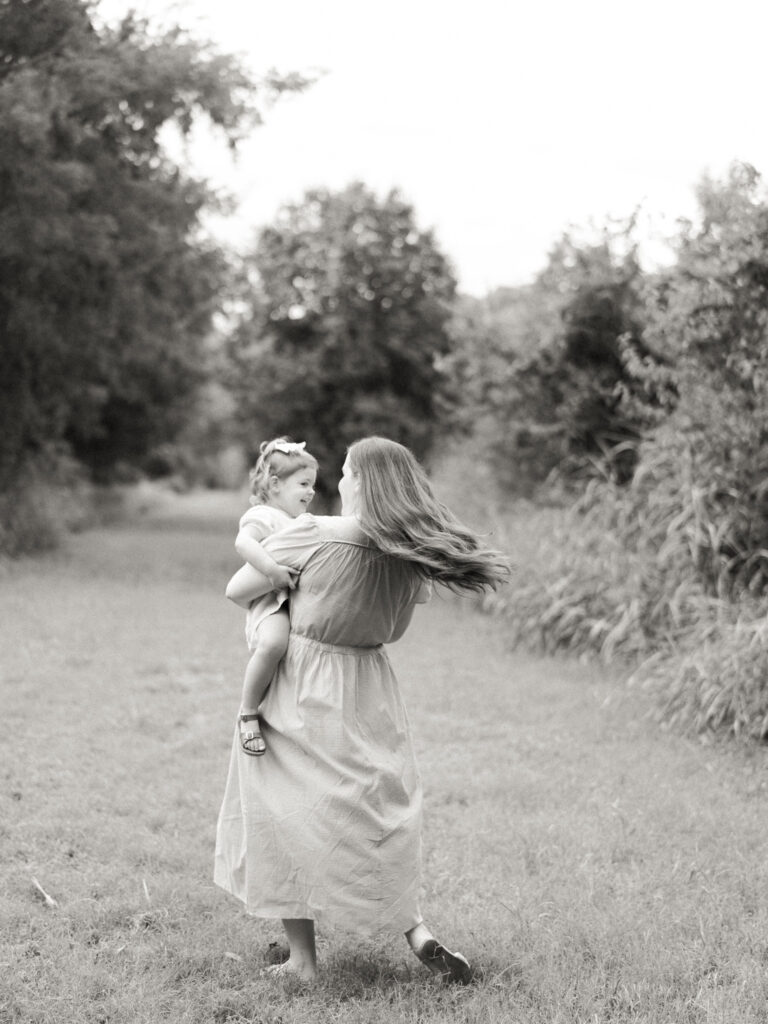 Monochromatic image of a mother holding a daughter while twirling in the grass. Images like this highlight the magic of a Little Rock Motherhood Session.