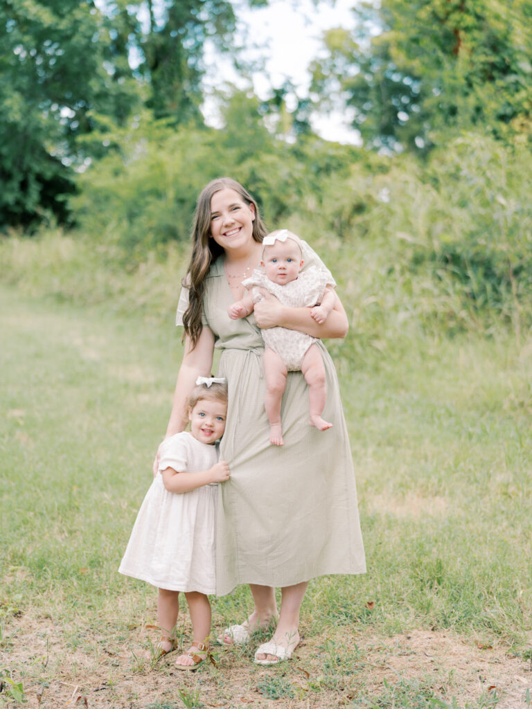 Mother in green dress smiles with her two young daughters embracing in a field of green grass and trees. Image derived from a Little Rock Motherhood Session.