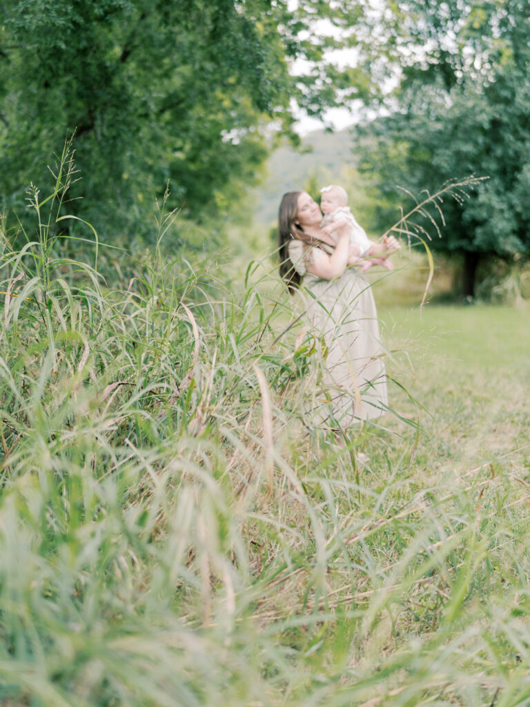 An out-of-focus mother snuggles her baby in the background, with a grassy foreground as a part of her Little Rock Motherhood Session.