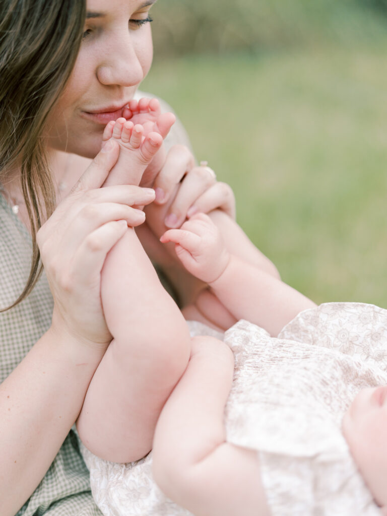 Brunette mother holds and kisses her out-of-frame 6 month old daughter's bare feet. Details like this are perfect for a Little Rock Motherhood Session.