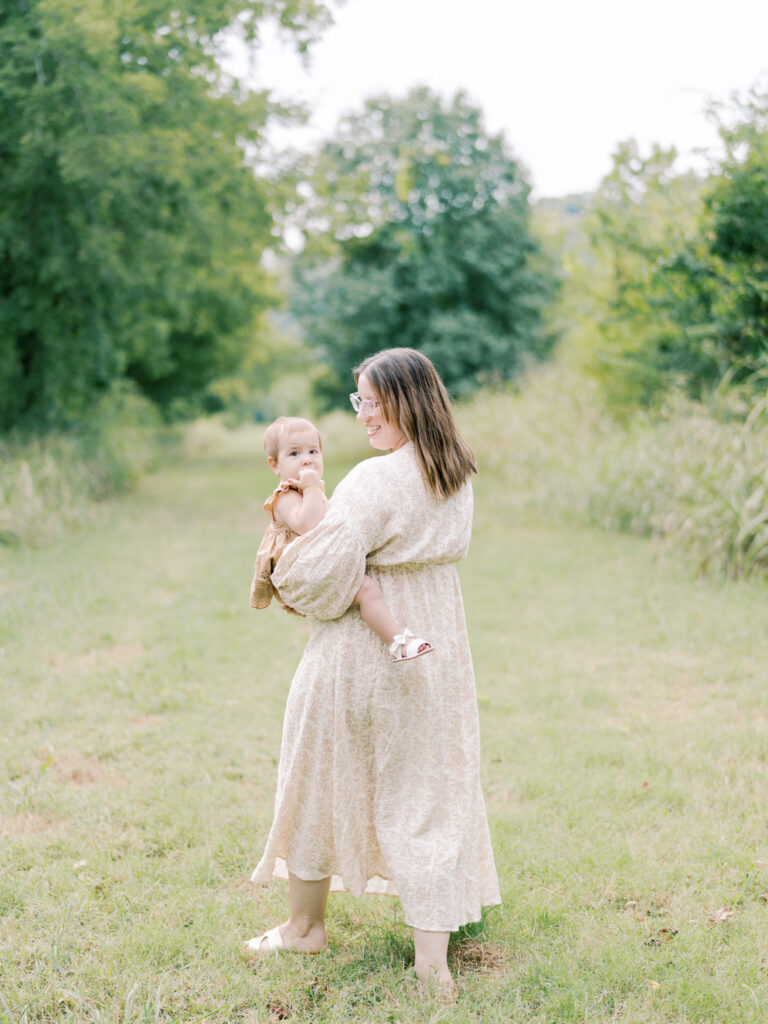 Brunette mother walking away while looking at the baby girl on her hip in a grassy, Little Rock field.