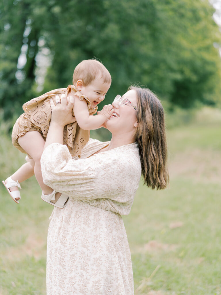 Brunette mother holding young daughter in the air while both giggle in a green, grassy field.