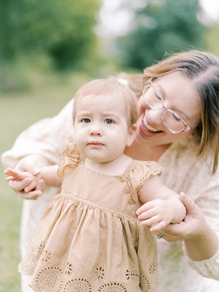 Brunette mother crouching down behind baby girl, holding her hands, while baby girl in brown dress looks at the camera.