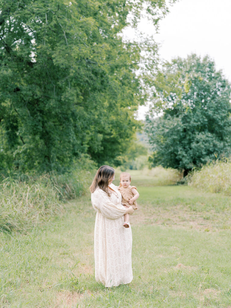 Mother in green, grassy field stands far away looking at the baby girl on her hip.