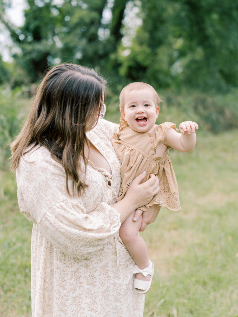 Mother looking at baby girl on her hip, while baby girl laughs at the camera, as a part of their Little Rock Motherhood Session.