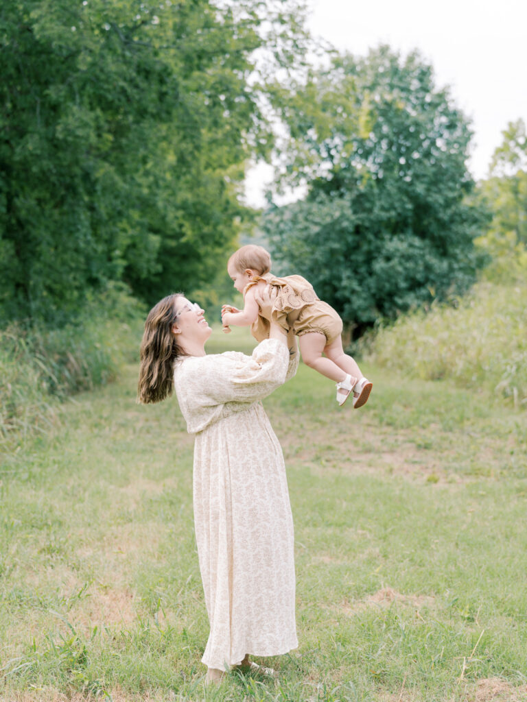 Brunette mother holding young daughter in the air while standing in a green, grassy field as a part of their Little Rock Motherhood Session.