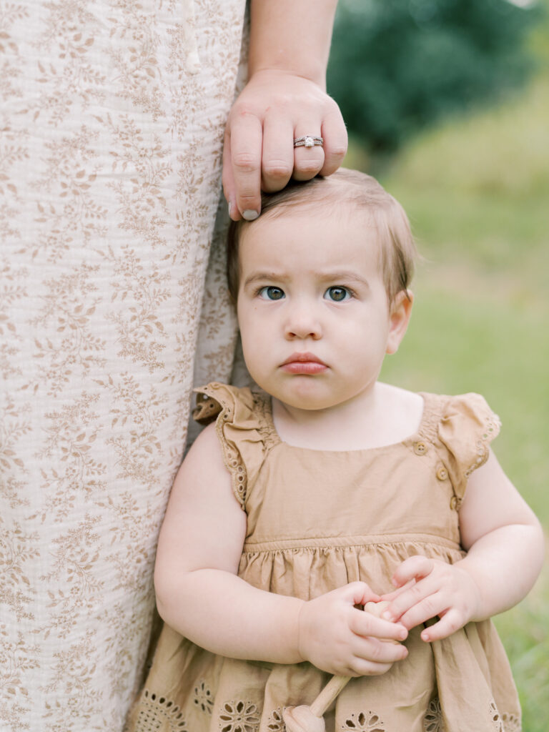 Somber baby girl in brown dress stands next to mother's leg while mother's hand reaches down to touch girl's hair, as a part of their Little Rock Motherhood Session.