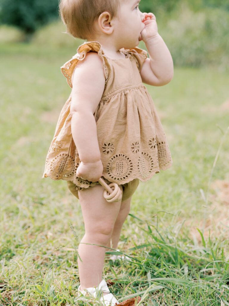 Side profile of a baby girl in a brown dress sucking her thumb with one hand and holding a wooden rattle in the other, while standing in a field of green grass.