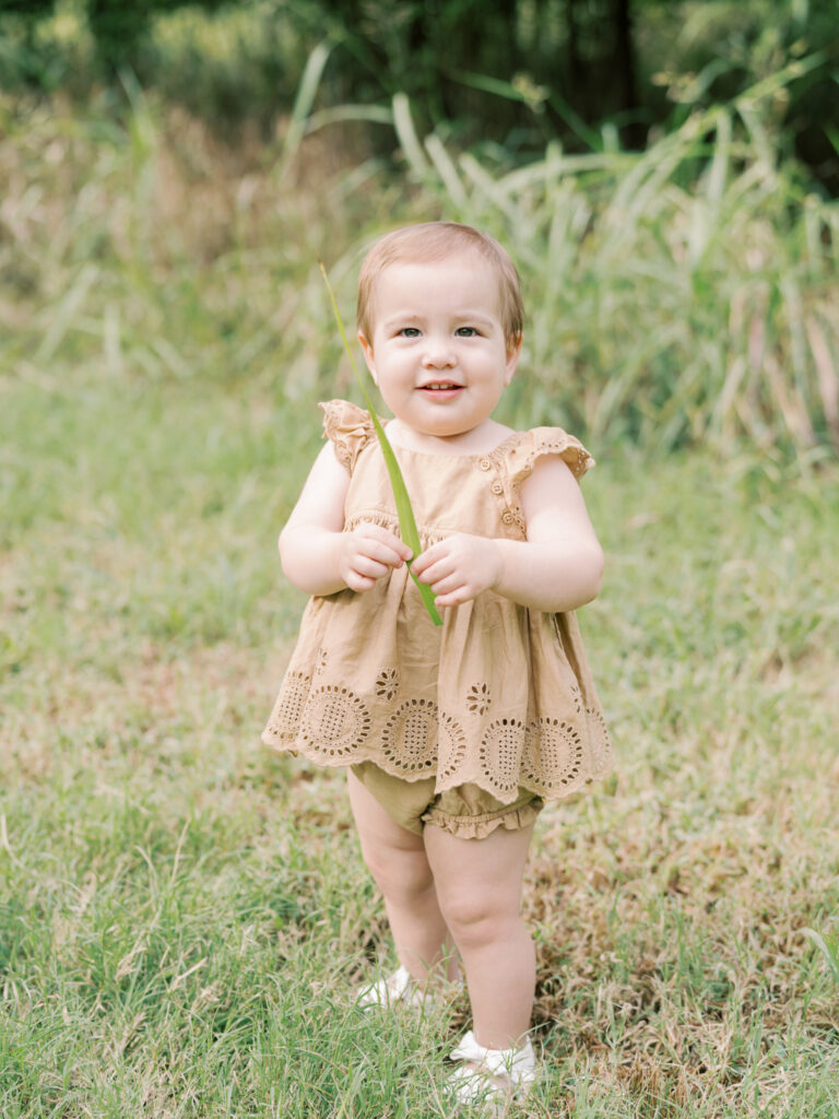 Baby girl in brown dress stands smiling while holding a blade of grass in a green, grassy field.