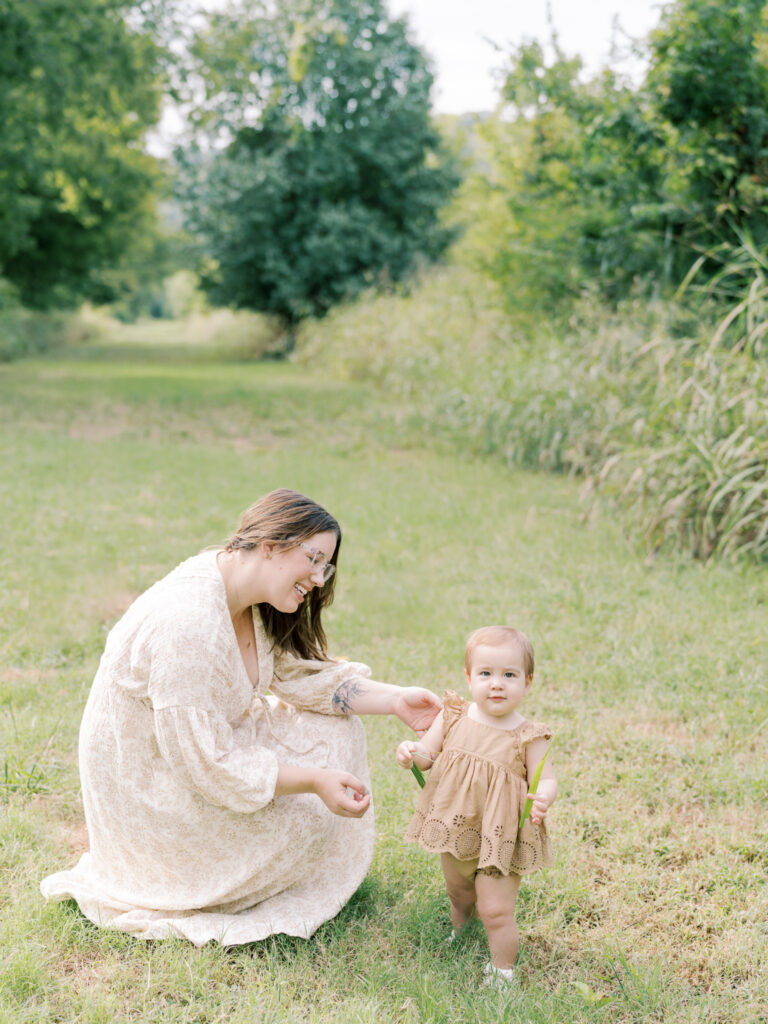 Brunette mother kneels in grass while looking at her standing baby girl in a brown dress, as a part of her Little Rock Motherhood Session.