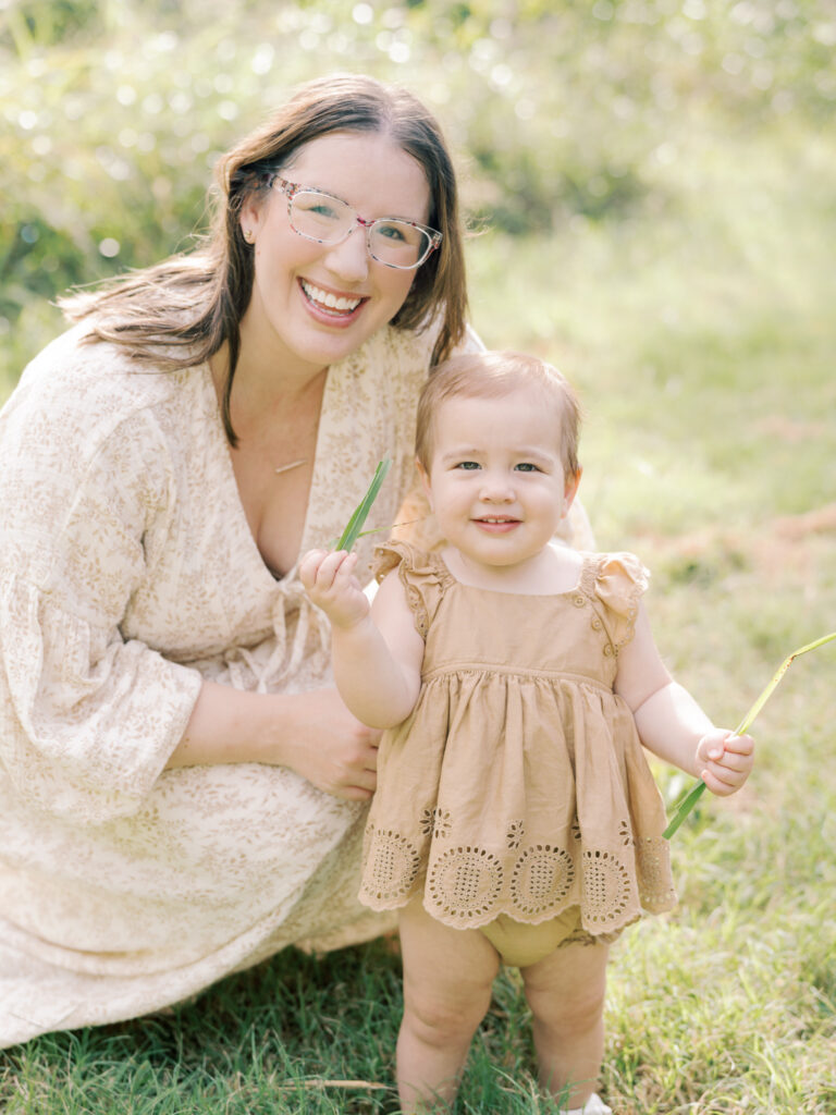 Crouching mother and standing baby girl daughter smile together in a field of green grass as a part of their Little Rock Motherhood Session.