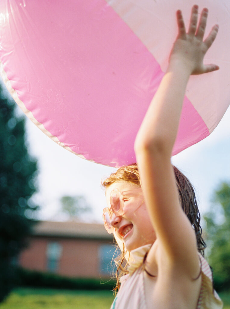 Young, smiling, red-headed girl in pink sunglasses faces the sun while holding huge, pink beachball above her head.