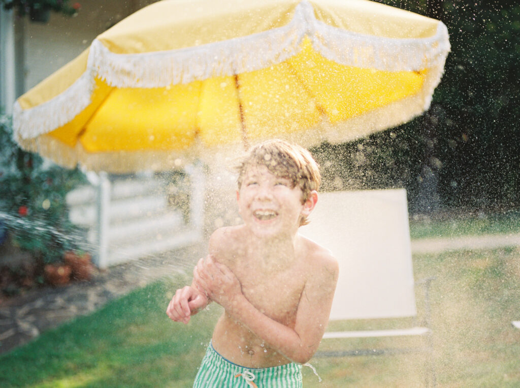 Boy standing in front of yellow yard umbrella gets sprayed by an off-camera water hose as he giggles.