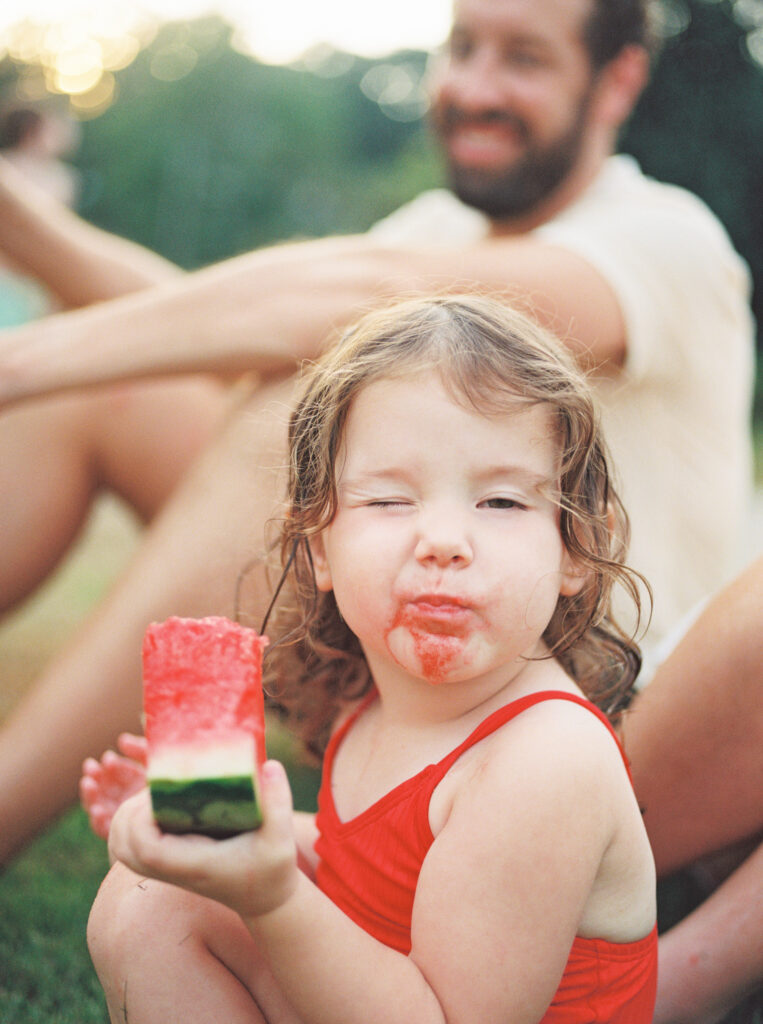 Toddler girl in red swimsuit, with watermelon dripping off her chin, winks at the camera.