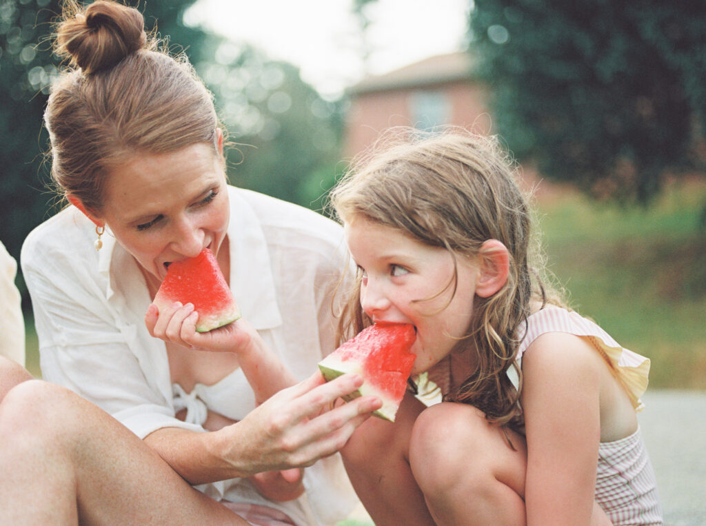 Mother and daughter sit in front yard and feed each other slices of watermelon.