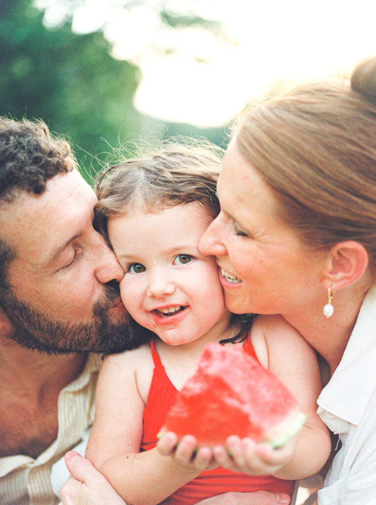 Mother and father kiss the cheeks of their toddler daughter as she eats watermelon.