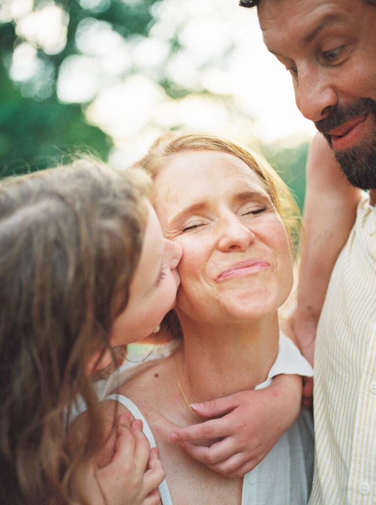 Young mother closes her eyes in joy as her daughter that's riding piggyback kisses her cheek.