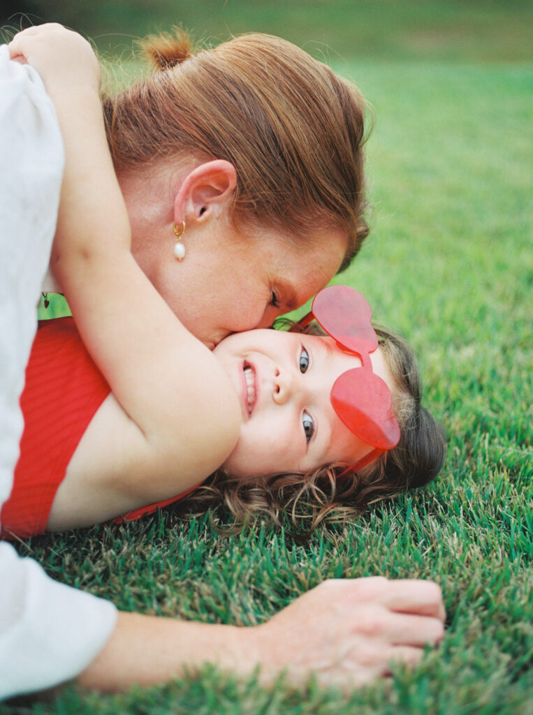 Mother and toddler daughter lie in green grass, as toddler in red heart sunglasses giggles when her mother kisses her.