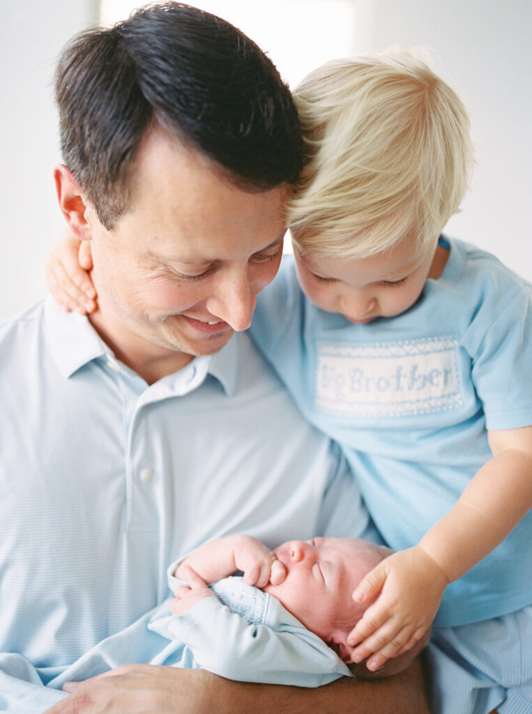 "Father holding newborn baby in soft natural light as toddler son embraces them both – film photo taken by Little Rock newborn photographer"