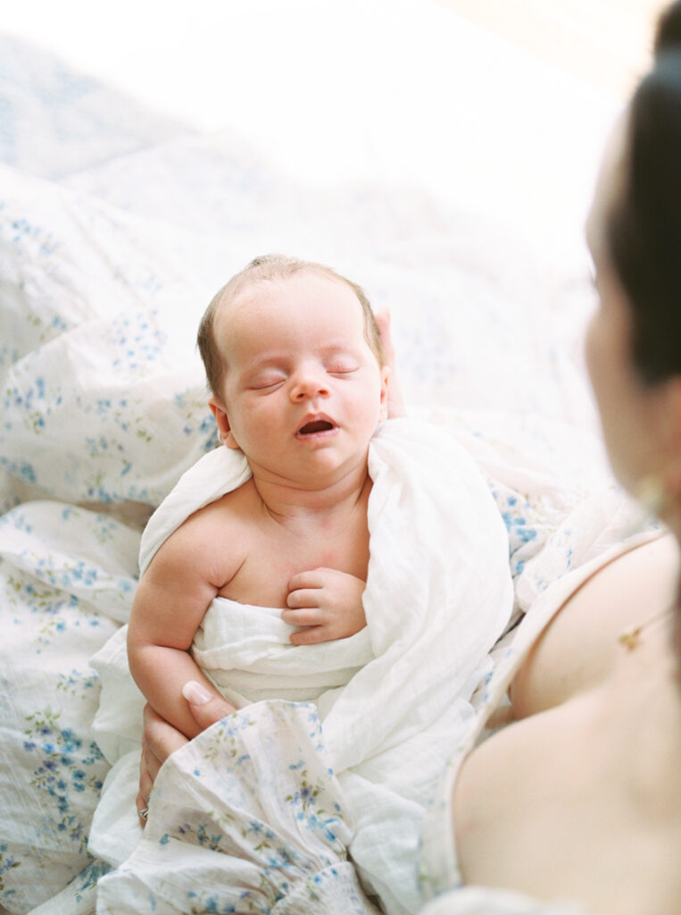 Baby boy in white muslin sleeps soundly in his mother's arms during their at-home newborn photography session, captured on film