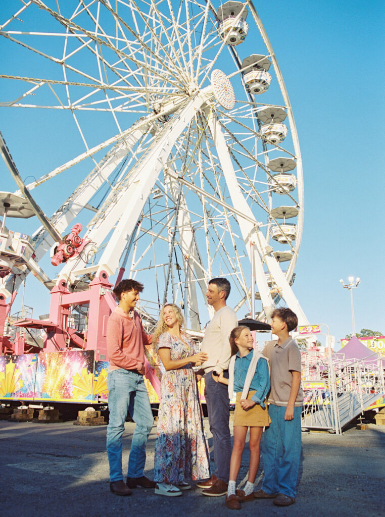 Family standing together in front of a Ferris wheel at the fair, smiling and laughing under bright blue skies — captured on film during a joyful family photography session focused on connection and togetherness.