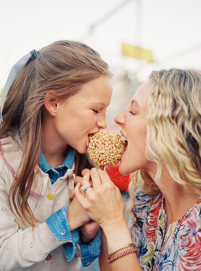 Mother and tween daughter sharing a caramel apple together at a fair, smiling and laughing during a playful moment — captured on film during a family photography session