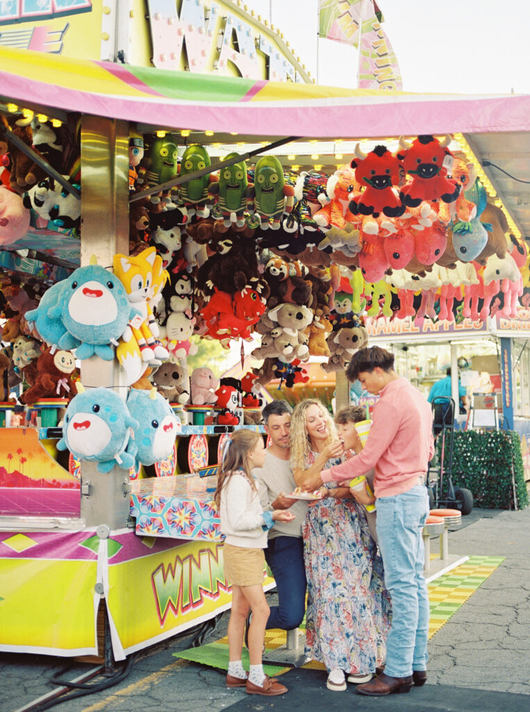 Family with tweens and teen enjoying a day together at a colorful fair game booth, surrounded by hanging stuffed animals and bright carnival lights — captured on film during a relaxed family photography session